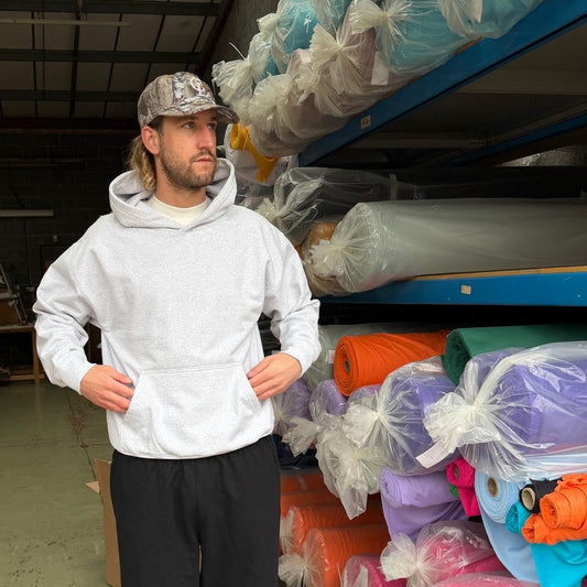 Man wearing a white hoodie and black pants standing in a warehouse with colorful fabric rolls.